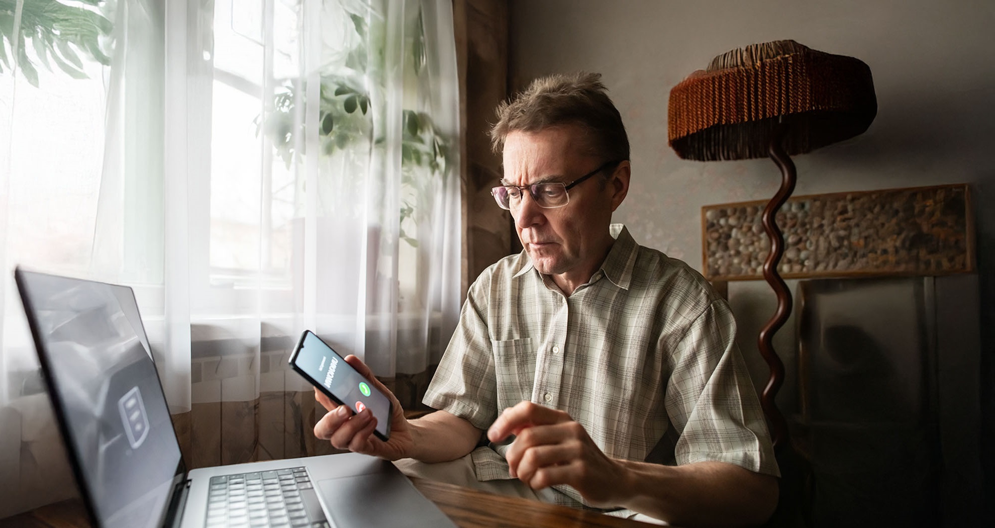 Older man sitting in front of a laptop and holding a phone, getting a call from an unknown number