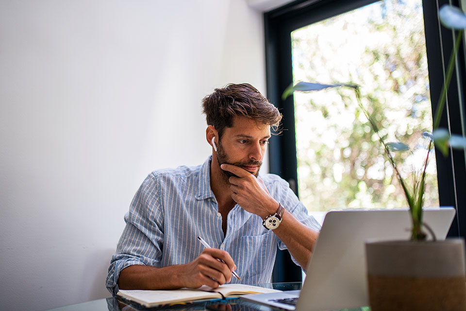 Man sitting in front of a laptop while writing in a notebook