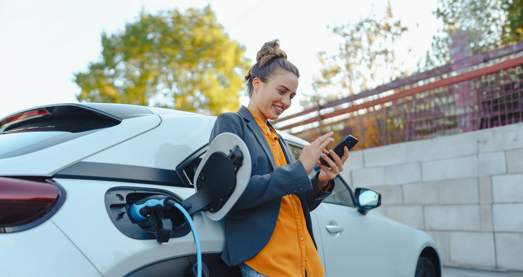 A woman leaning against her charging electric car, checking out AIB's car insurance deals on her phone.