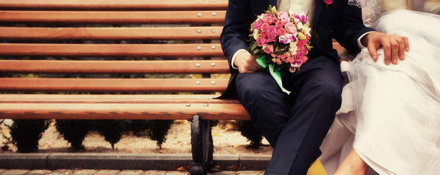 A married couple sitting on a bench in their wedding clothes.