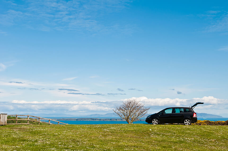 A black car with its boot open parked on grass near the coast.