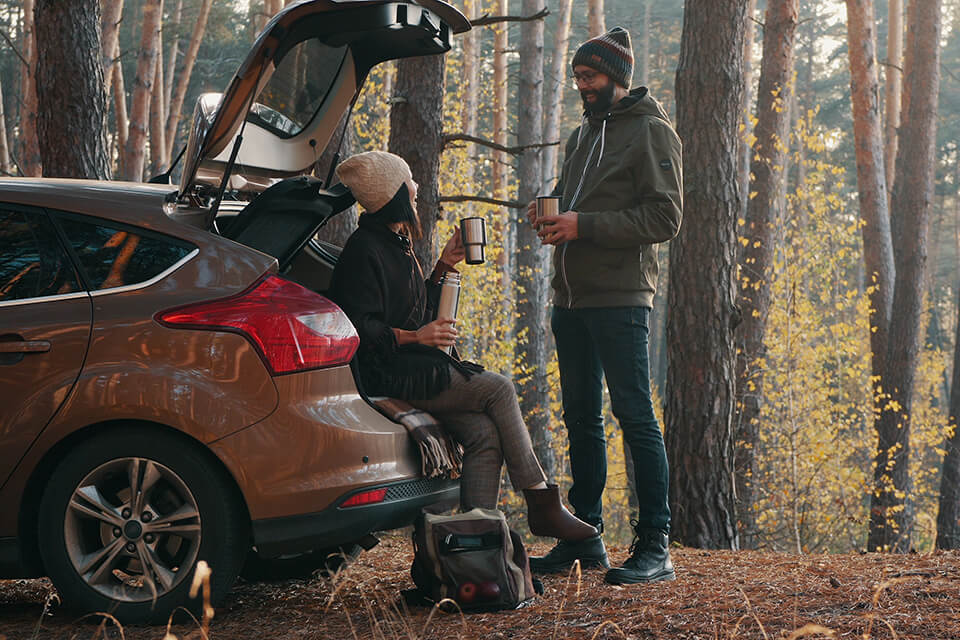 A woman sitting on the bumper of a car parked in forrest sharing tea with a man standing opposite of her.