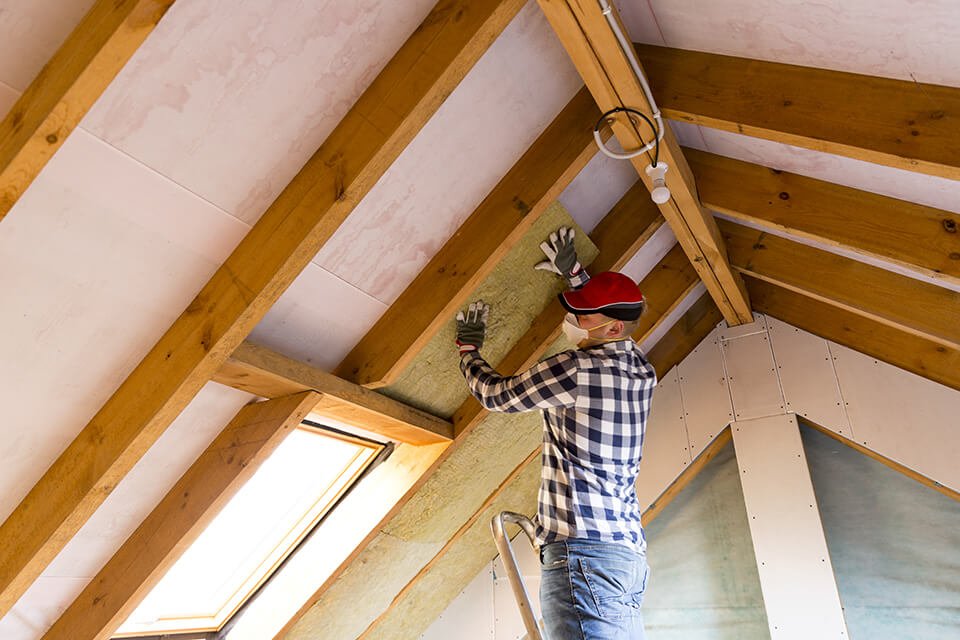 A man wearing a mask installing insulation above a window in the attic.