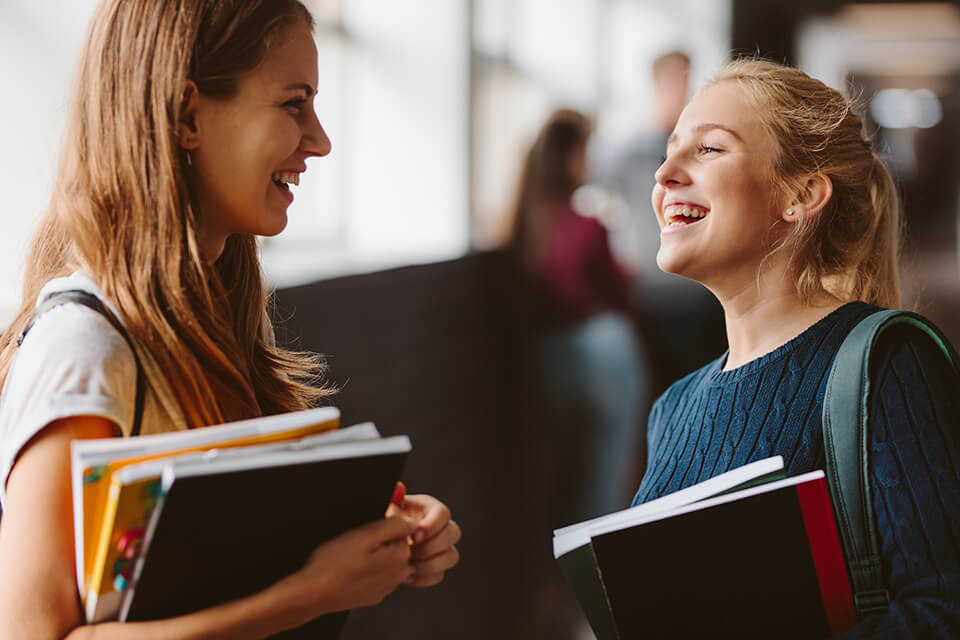 Two female students holding their books while chatting in the hallway.
