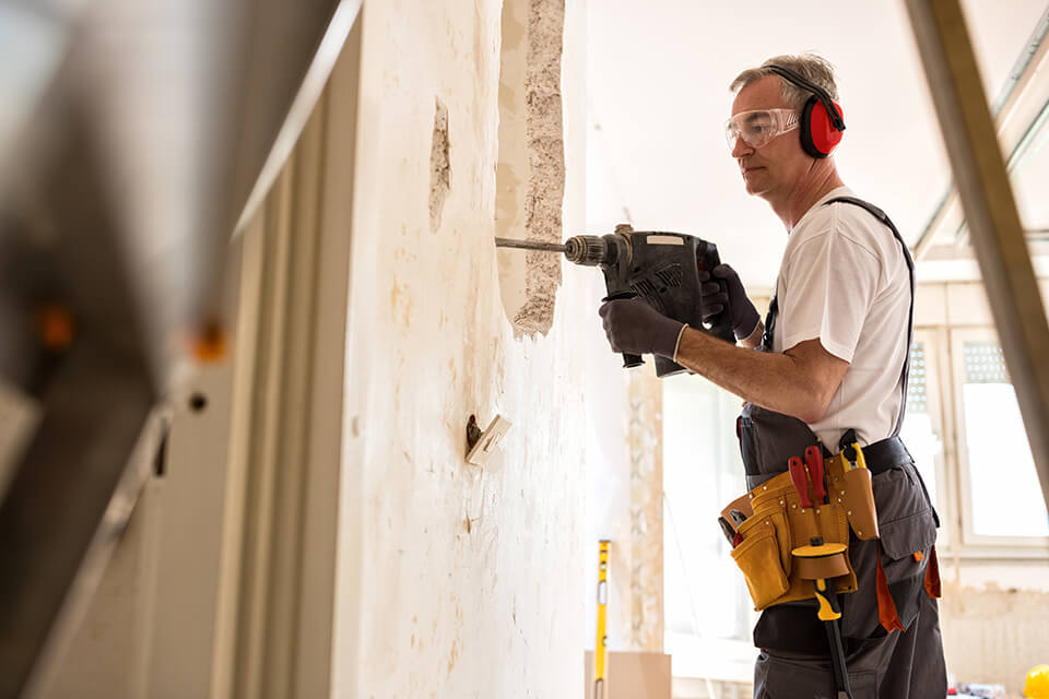 A construction worker drilling a hole in a wall inside a house.