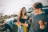 A couple standing beside their car eating lunch by the beach.
