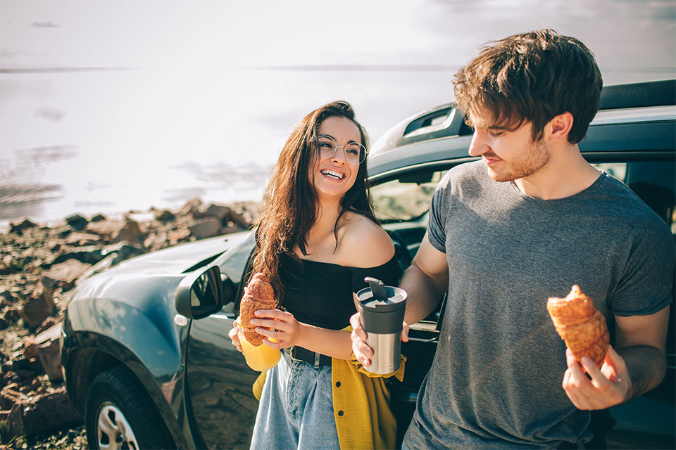 A couple standing beside their car eating lunch by the beach.