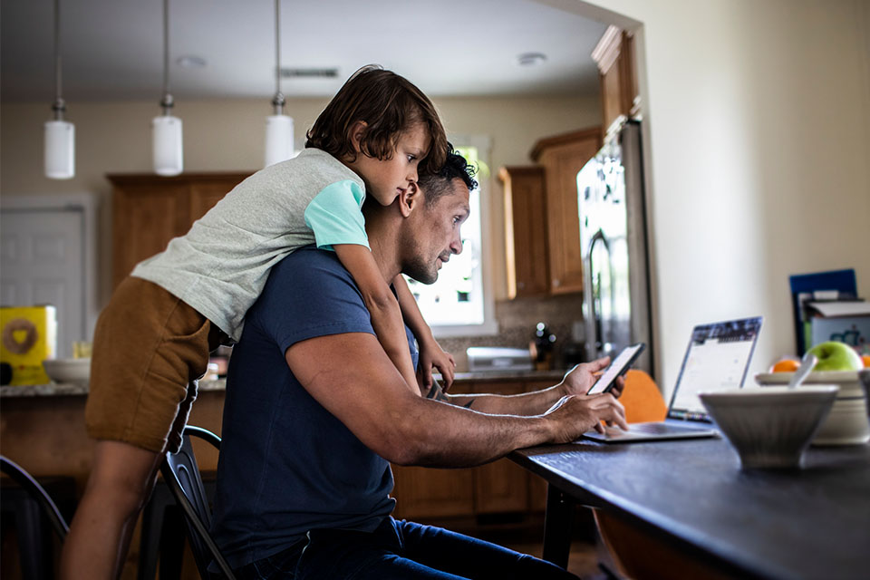 Man sitting at a desk looking at his phone with his son leaning over his shoulders.