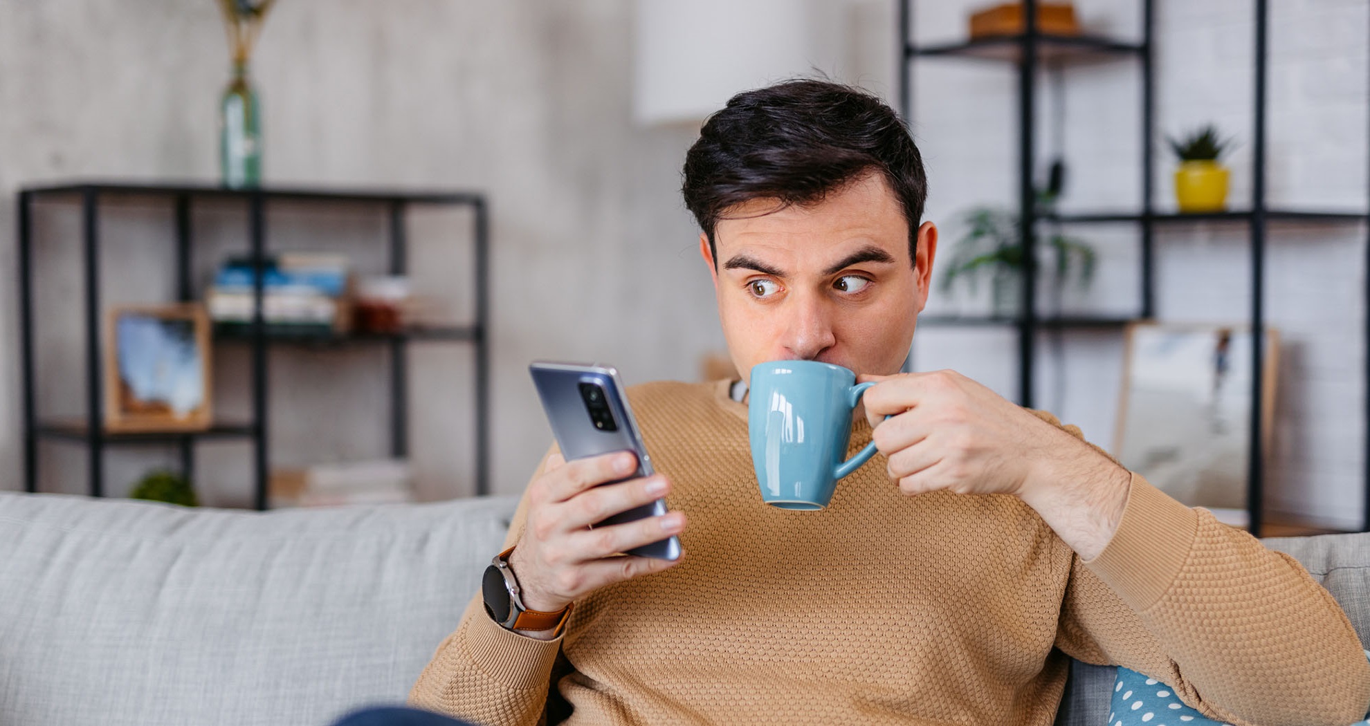 Man sitting on a sofa drinking from a mug looking at his phone.