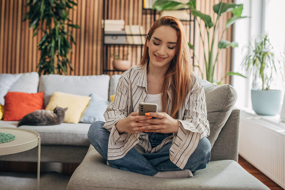 Woman sitting cross legged on a sofa smiling at her phone.